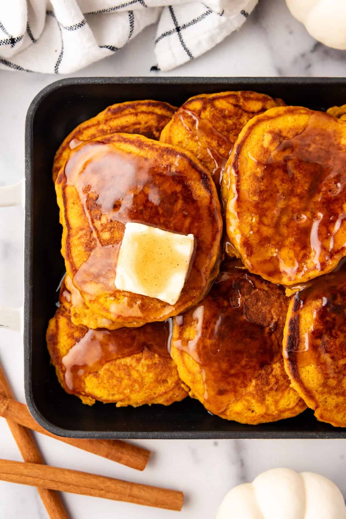 An overhead image of pumpkin pancakes with cinnamon syrup and butter on top next to cinnamon sticks and butter.