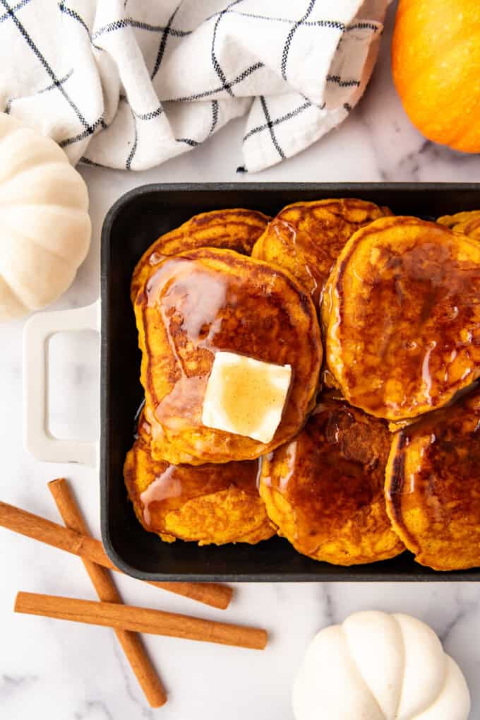 An overhead image of pumpkin pancakes with cinnamon syrup and butter on top next to cinnamon sticks and butter.