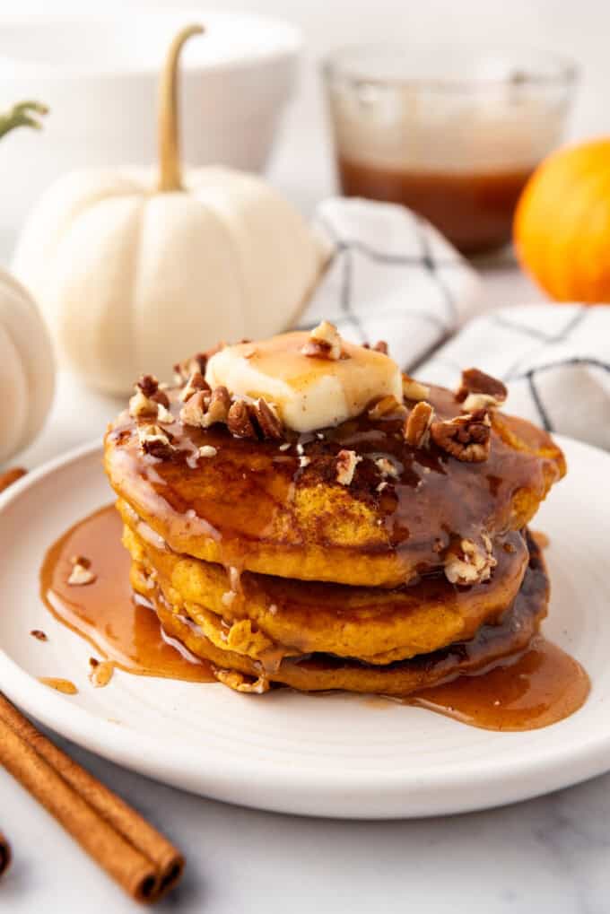 A stack of pumpkin pancakes on a white plate with pumpkins and syrup in the background.