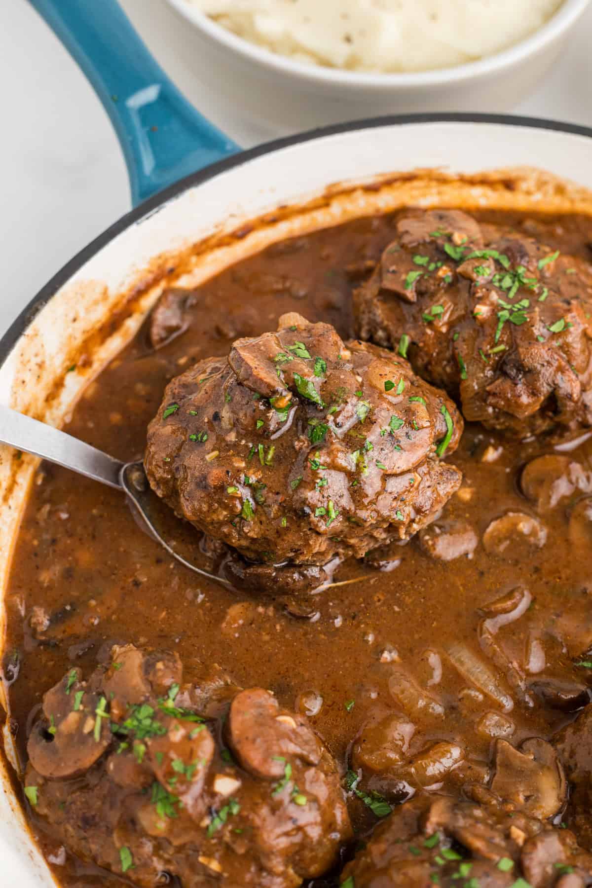 A spoon lifting a salisbury steak out of a pan.