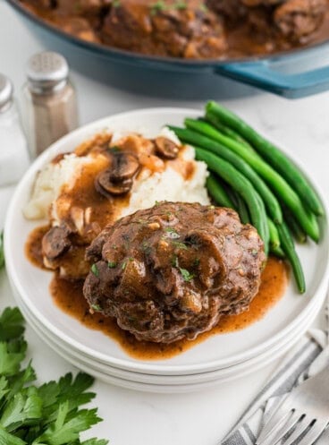 A side view of a white plate with a salisbury steak on it with mashed potatoes and green beans.