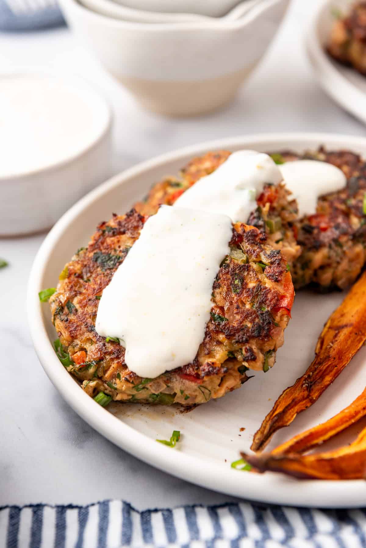 Salmon cakes topped with a white sauce, on a white plate with a side of sweet potato fries.