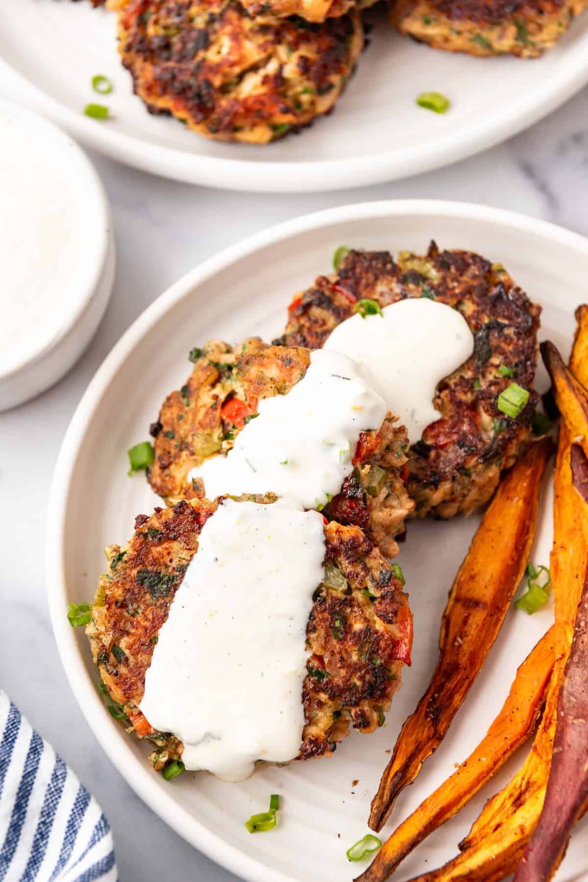 Salmon cakes topped with a white sauce, on a white plate with a side of sweet potato fries.