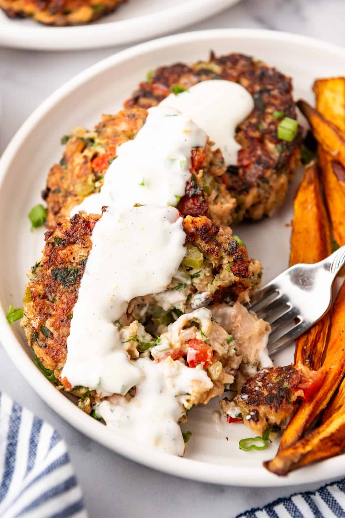 Salmon cakes topped with a white sauce, on a white plate with a side of sweet potato fries.