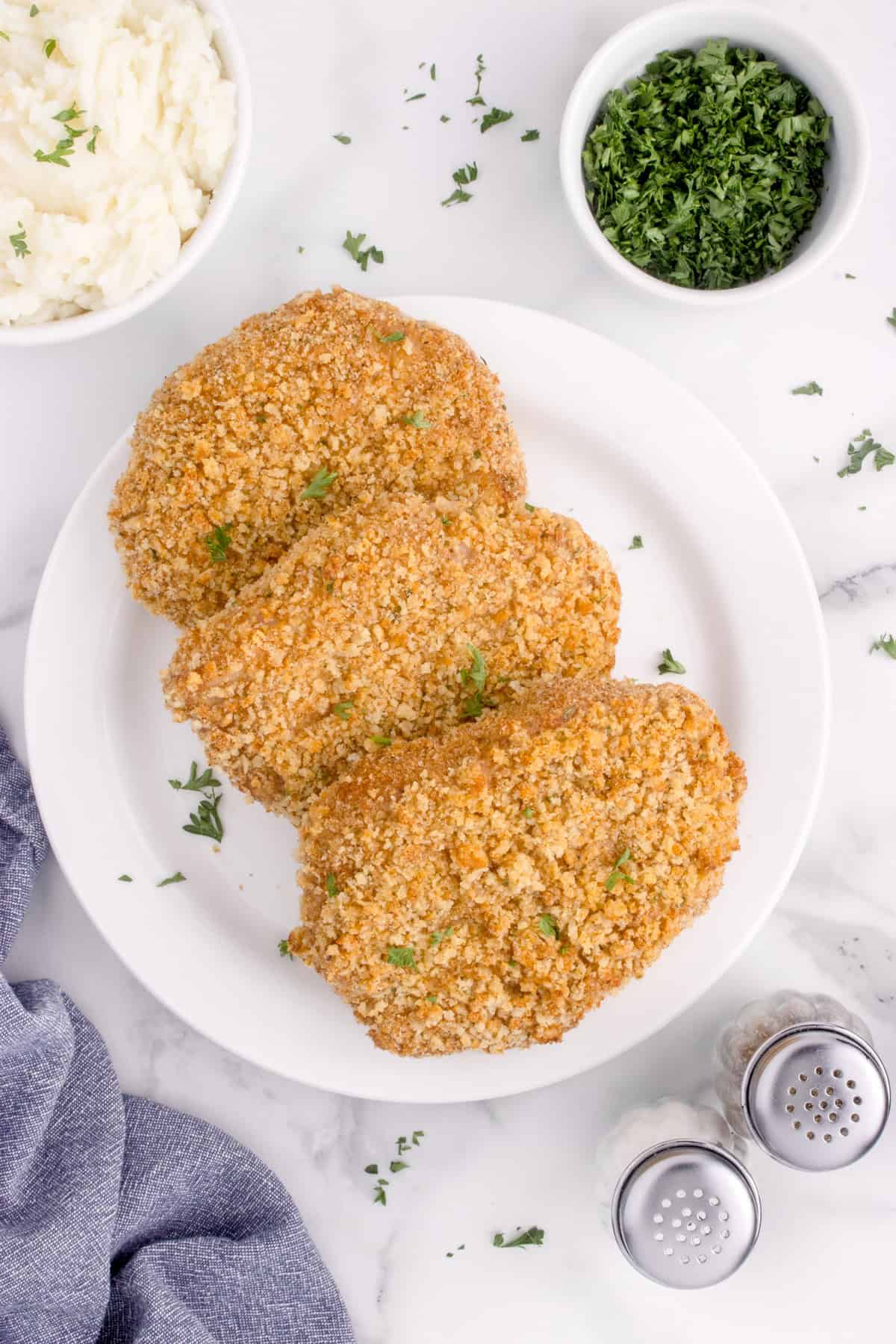 An overhead image of cooked shake n bake pork chops on a white plate next to a bowl of mashed potatoes.