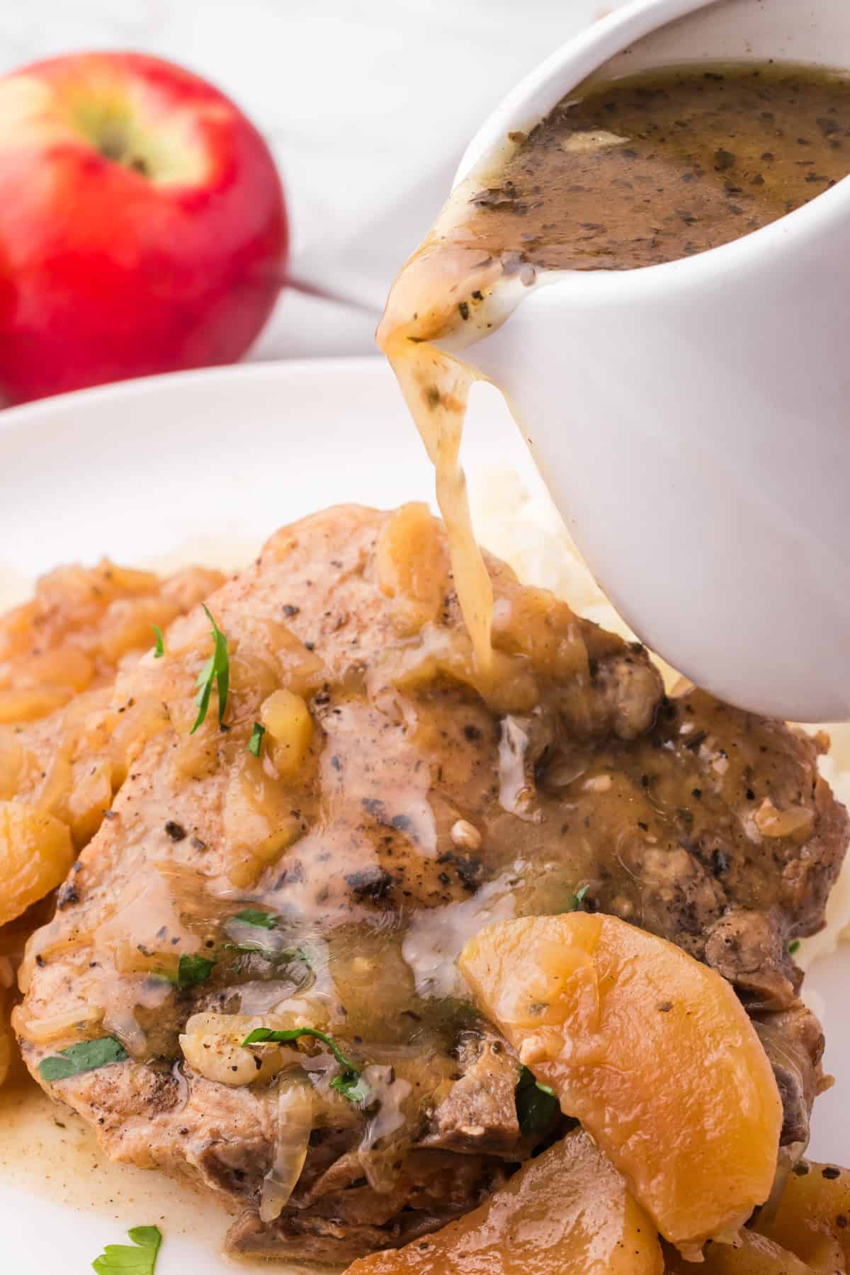 Sauce being poured over slow cooked pork chops on a plate with cooked apples.