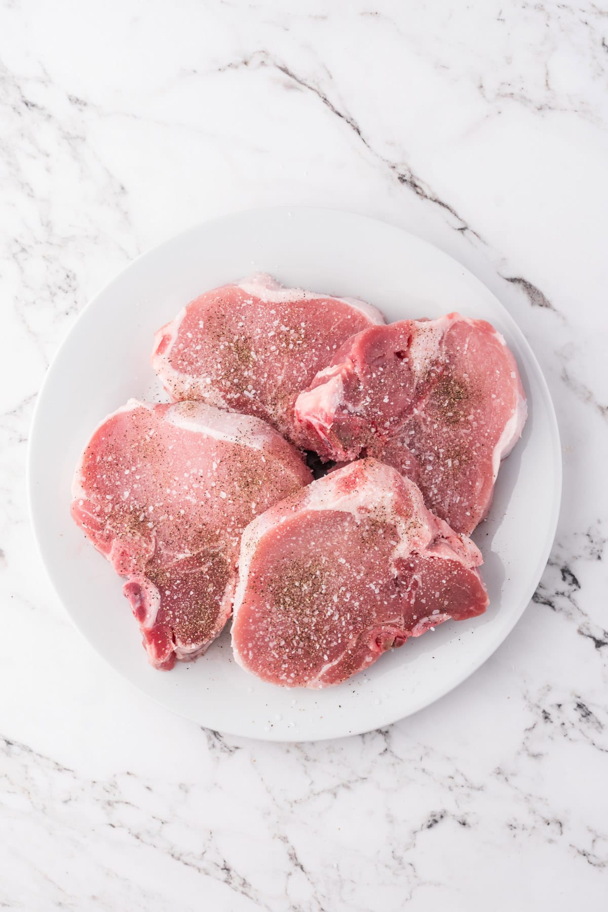 Seasoned pork chops on a white plate before cooking.