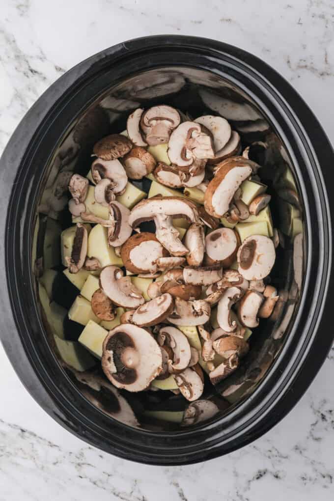 Adding sliced mushrooms and potatoes to a crock pot.