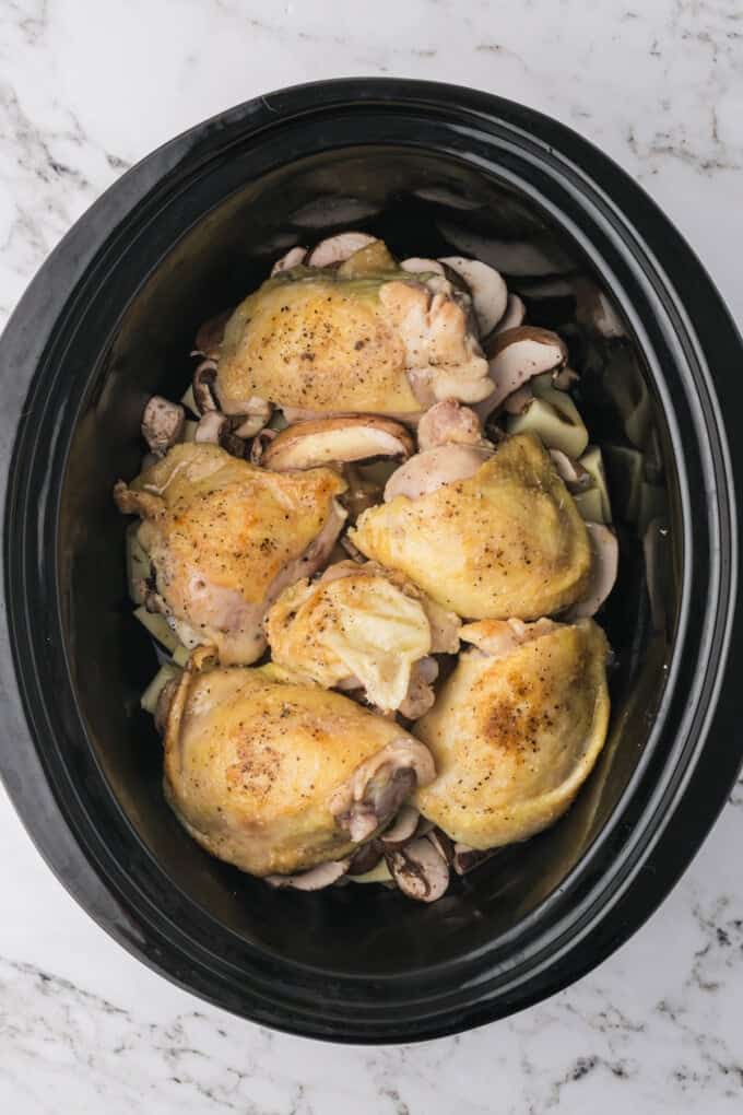 Adding seared chicken thighs to a crock pot.