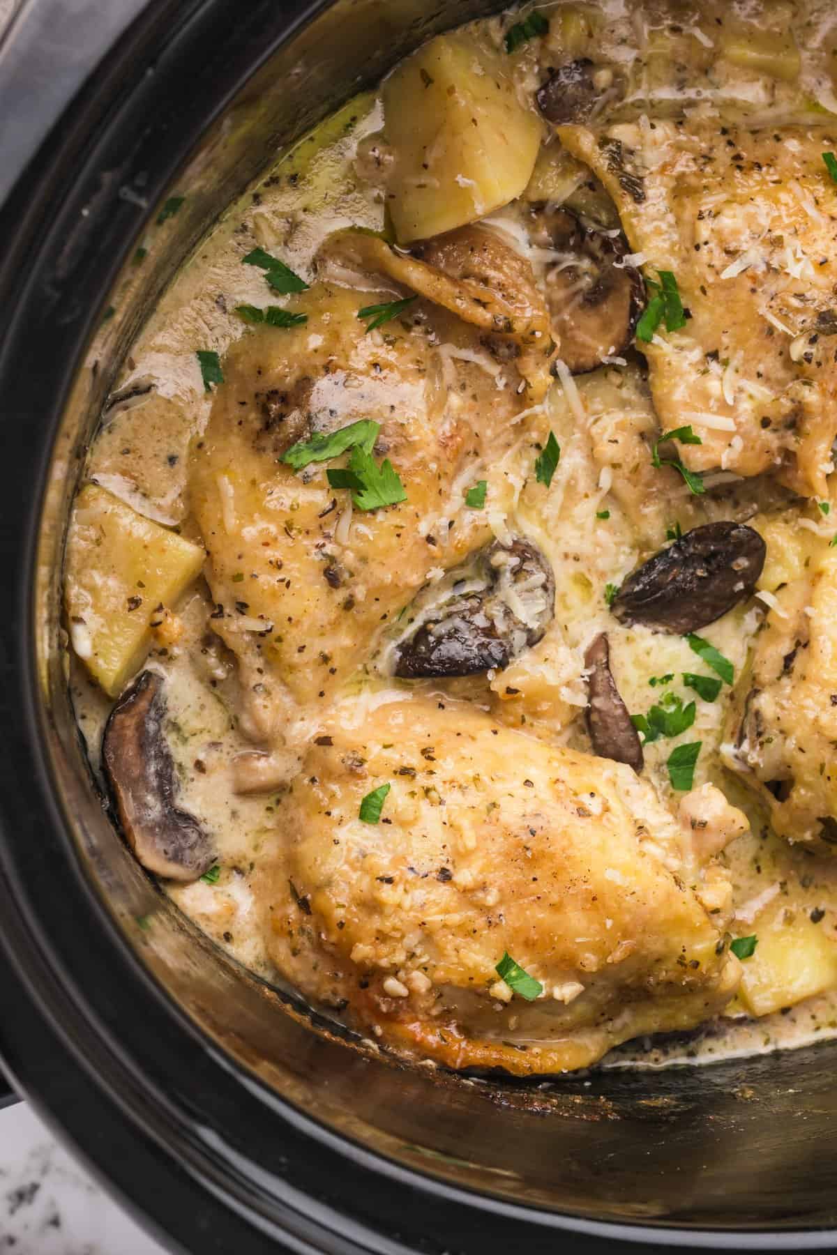 An overhead image of chicken thighs in a crock pot with a garlic cream sauce, mushrooms, and potatoes.