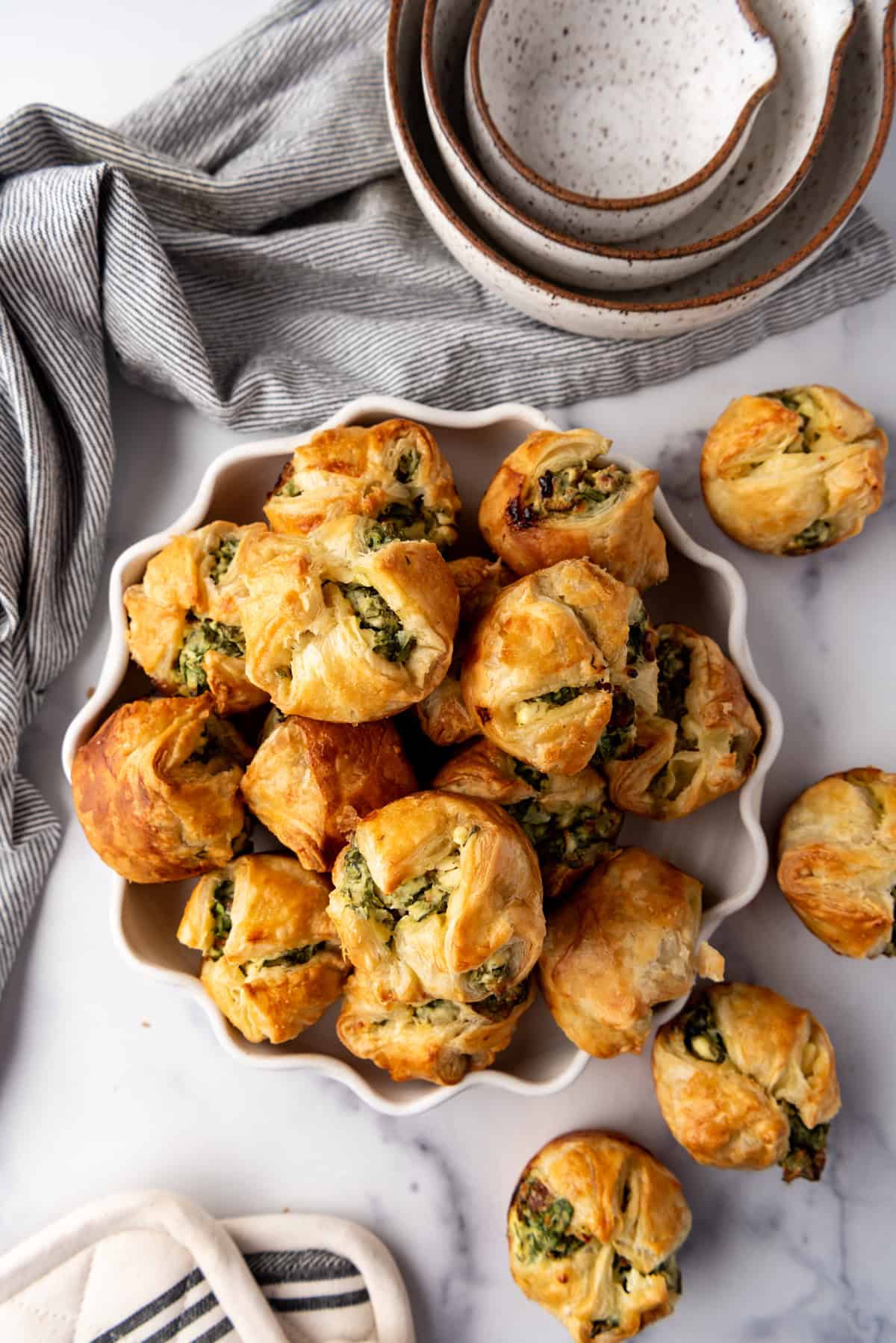 An overhead image of a bowl of spinach puffs next to more spinach puffs, mixing bowls, and a cloth napkin.