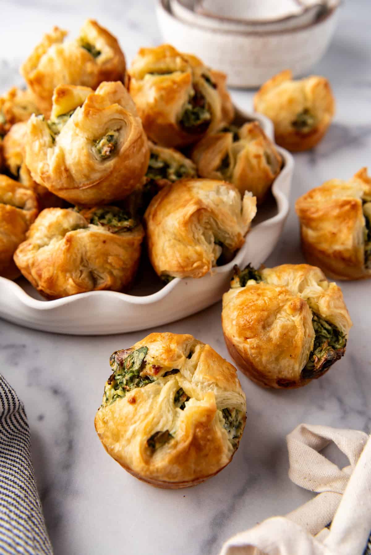 Spinach puffs on a marble surface in front of more of them in a bowl.