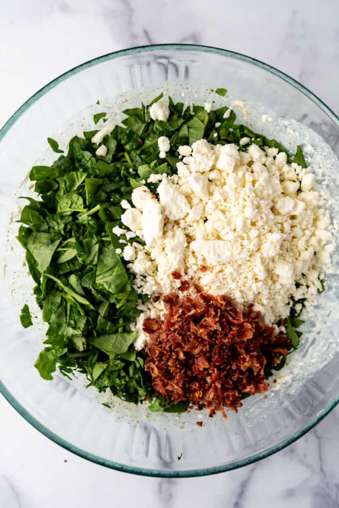 Adding chopped fresh spinach, feta cheese, and crumbled bacon bits to a large mixing bowl.