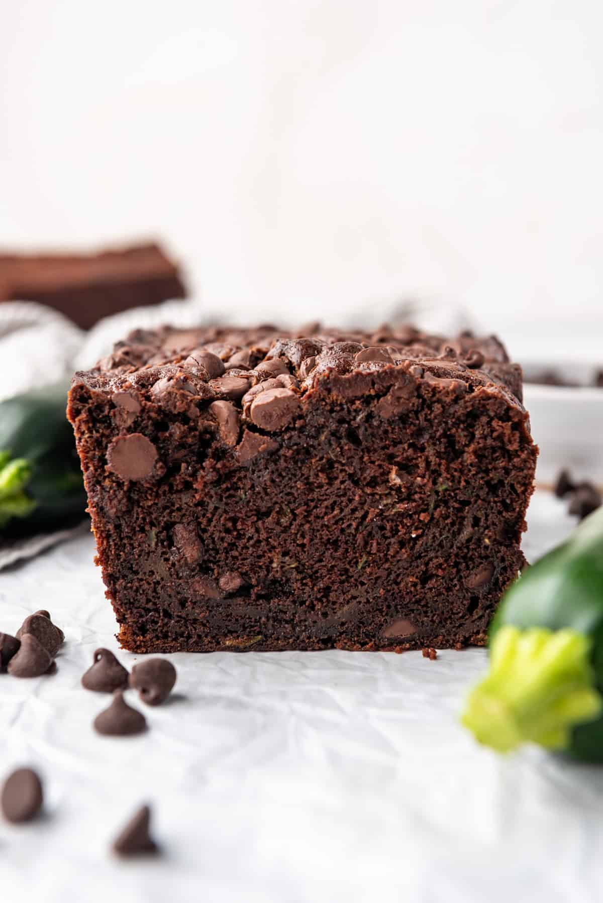 A loaf of chocolate zucchini bread on a white surface with zucchini and chocolate chips around it.