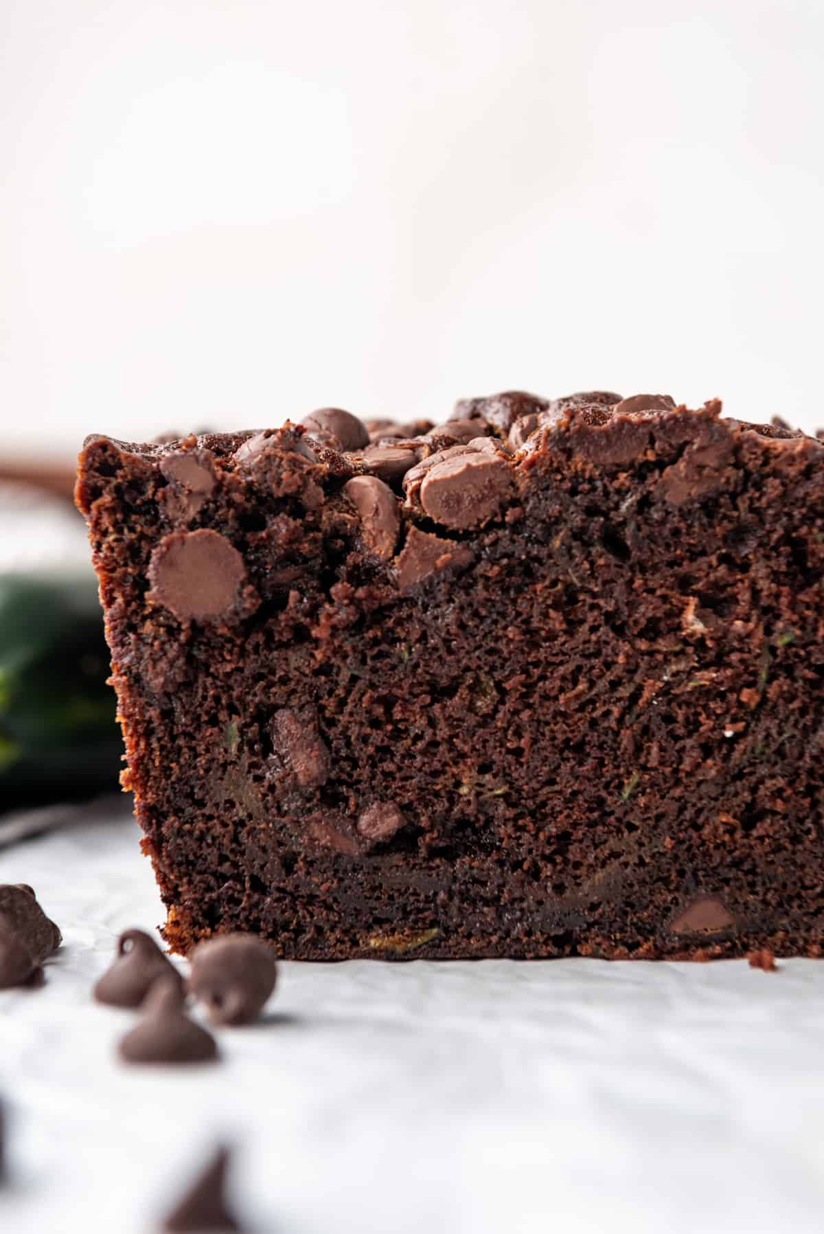 A loaf of chocolate zucchini bread on a white surface with zucchini and chocolate chips around it.