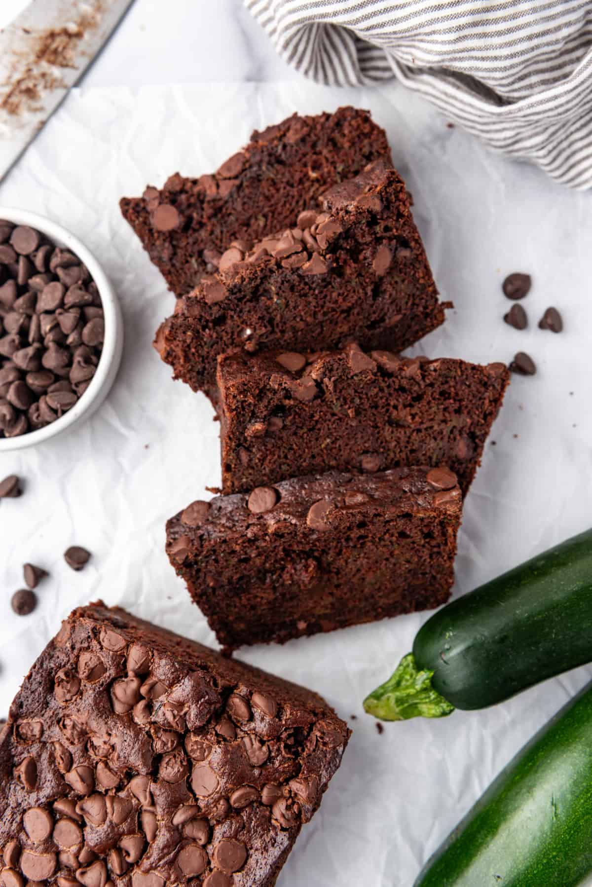 Top view of slices of chocolate zucchini bread on a white surface with zucchini and chocolate chips around them.