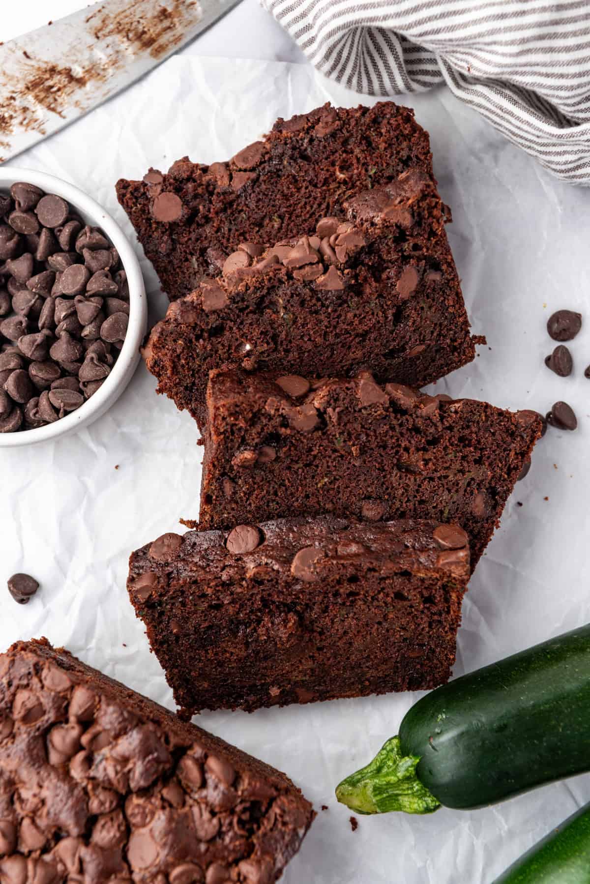 Top view of slices of chocolate zucchini bread on a white surface with zucchini and chocolate chips around them.