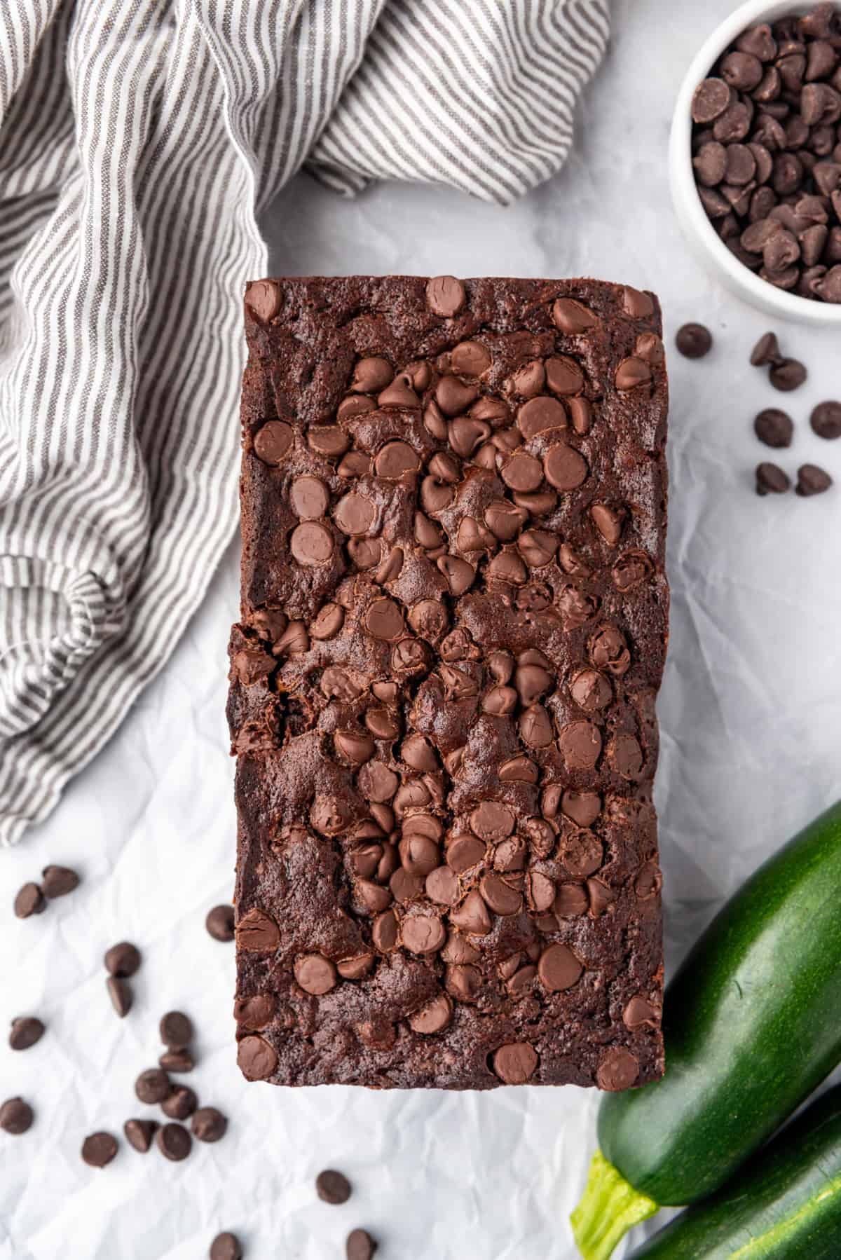 Top view of a loaf of chocolate zucchini bread on a white surface with zucchini and chocolate chips around it.