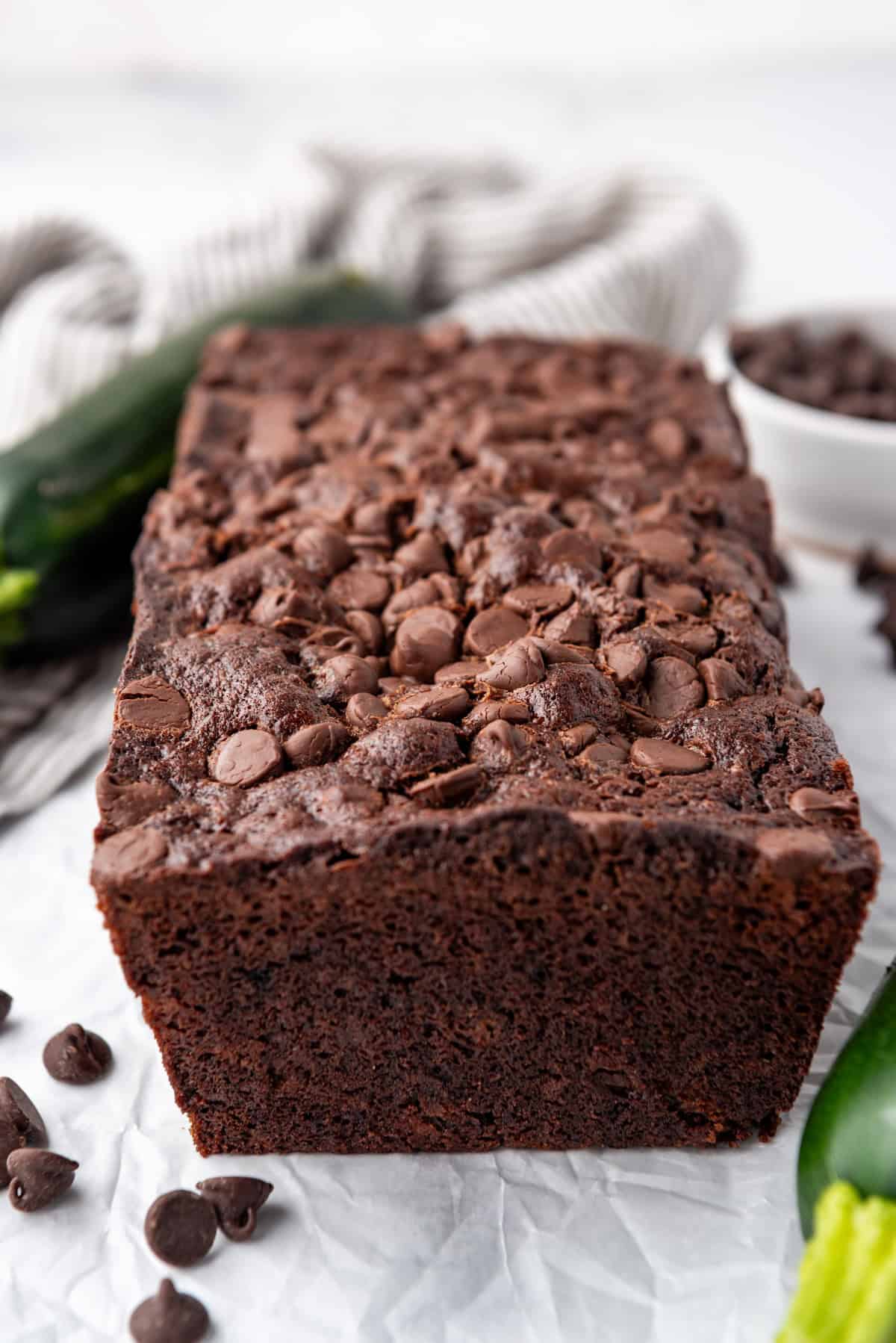 A loaf of chocolate zucchini bread on a white surface with zucchini and chocolate chips around it.