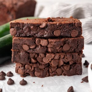 Slices of chocolate zucchini bread stacked on a white surface with zucchini and chocolate chips around them.