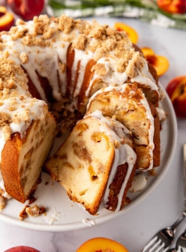 An image of a sliced peach cobbler pound cake on a white cake plate with fresh peaches and a green and white checkered cloth nearby.