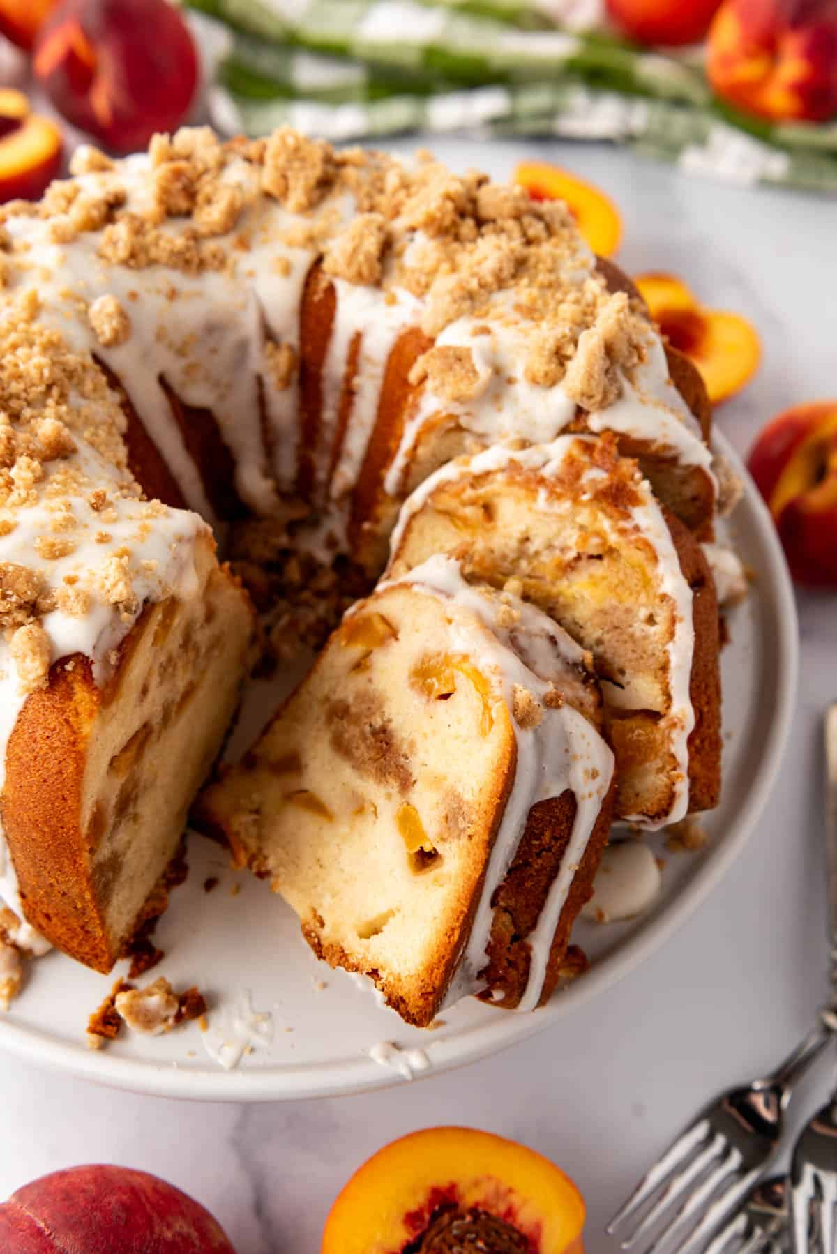 An image of a sliced peach cobbler pound cake on a white cake plate with fresh peaches and a green and white checkered cloth nearby.