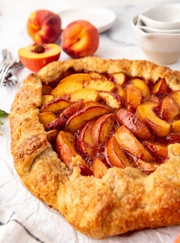 A side angle of a peach galette in front of fresh peaches and measuring cups.