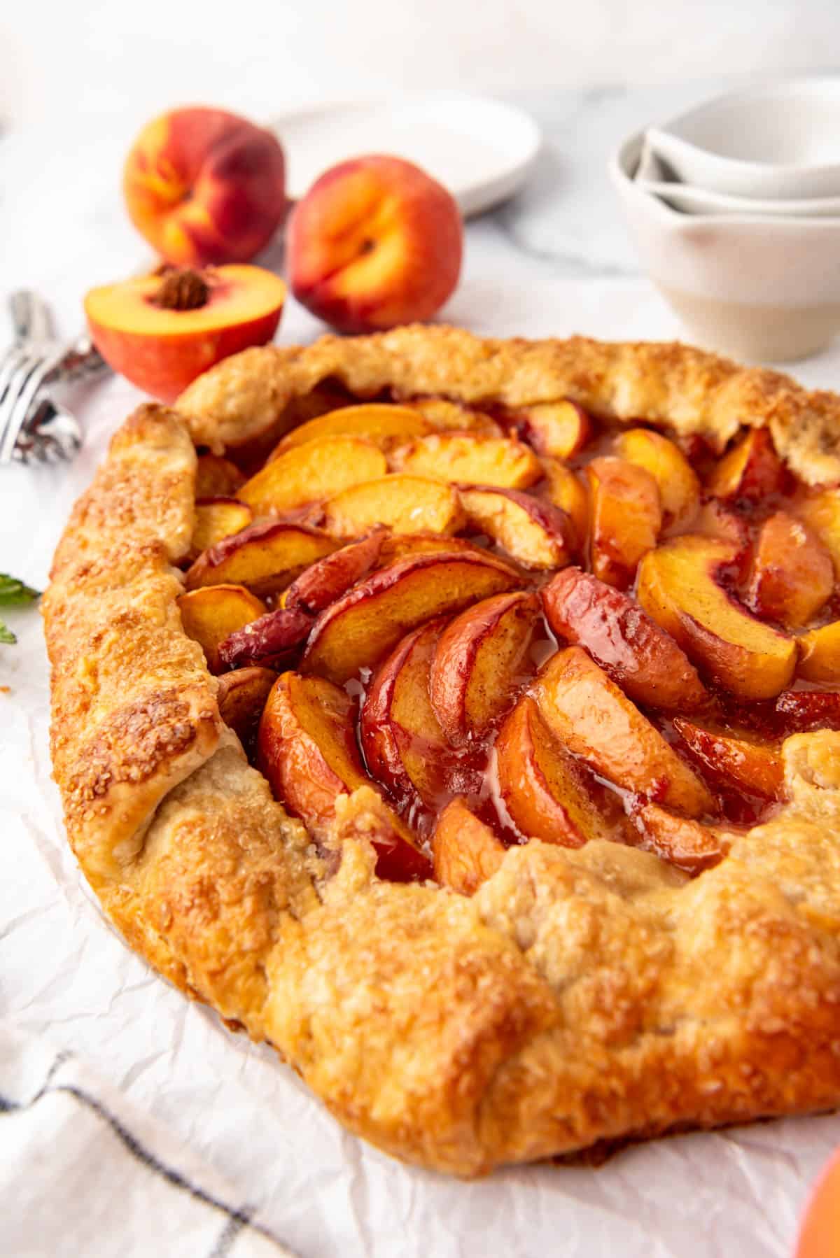 A side angle of a peach galette in front of fresh peaches and measuring cups.