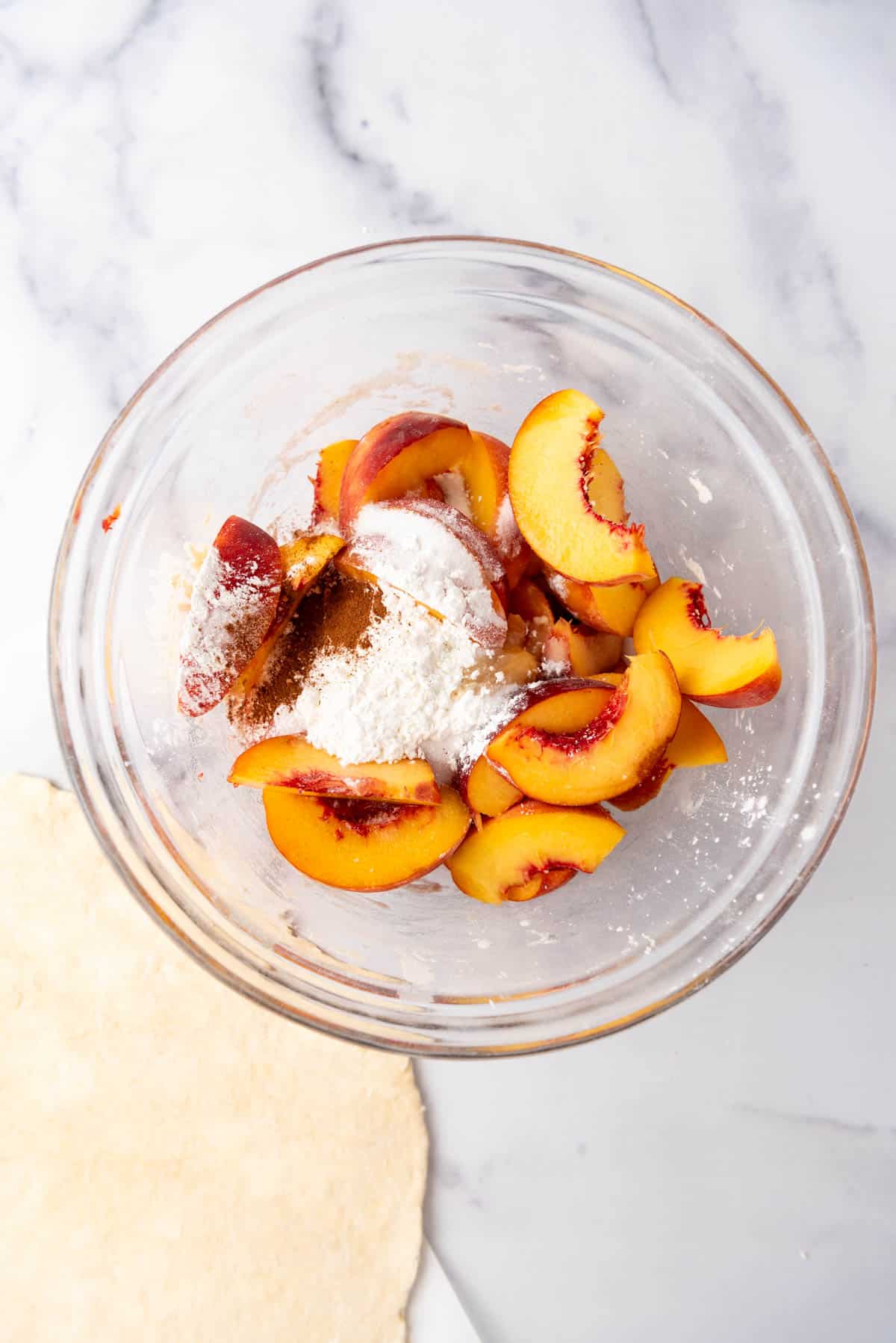 Combining sliced peaches, sugar, cornstarch, and cinnamon in a glass mixing bowl to make a galette.