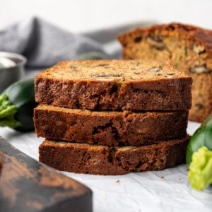 Slices of Zucchini Bread stacked on top of each other next to a wooden chopping board on a white surface.