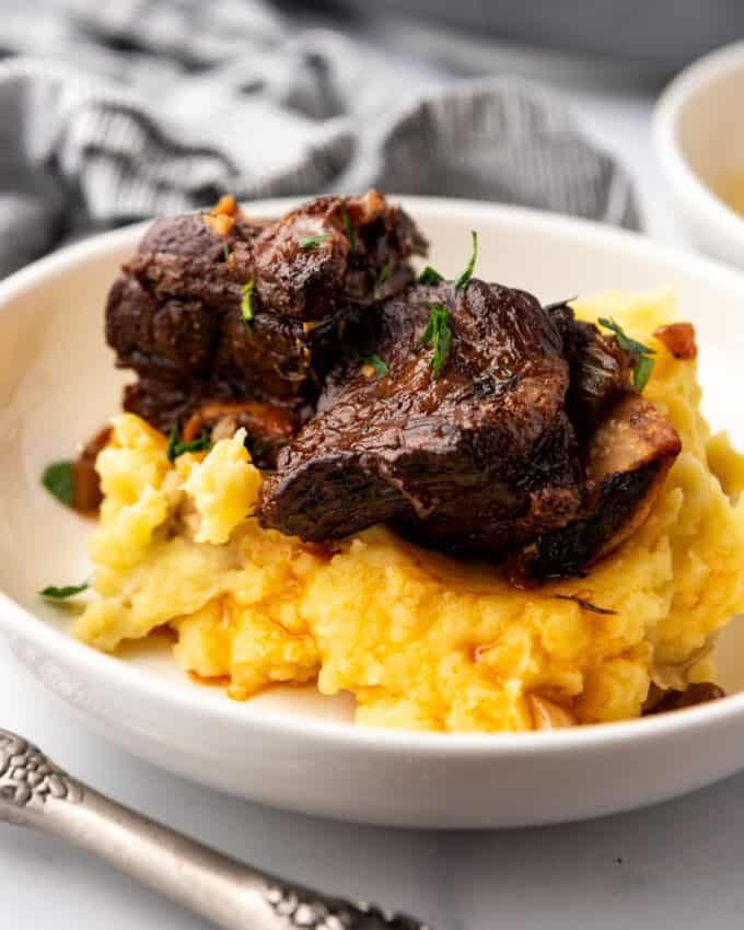 Close up image of a serving of Braised Short Ribs on a bed of mashed potatoes, in a white plate on a white countertop.