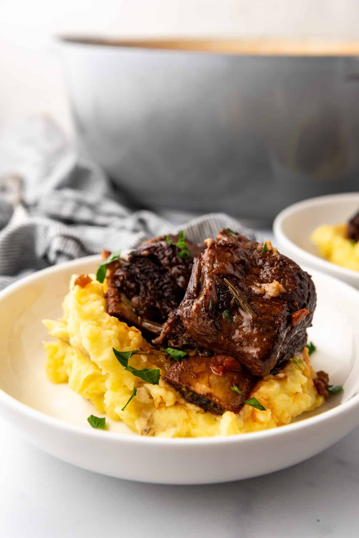 Close up image of a serving of Braised Short Ribs on a bed of mashed potatoes, in a white plate on a white countertop.