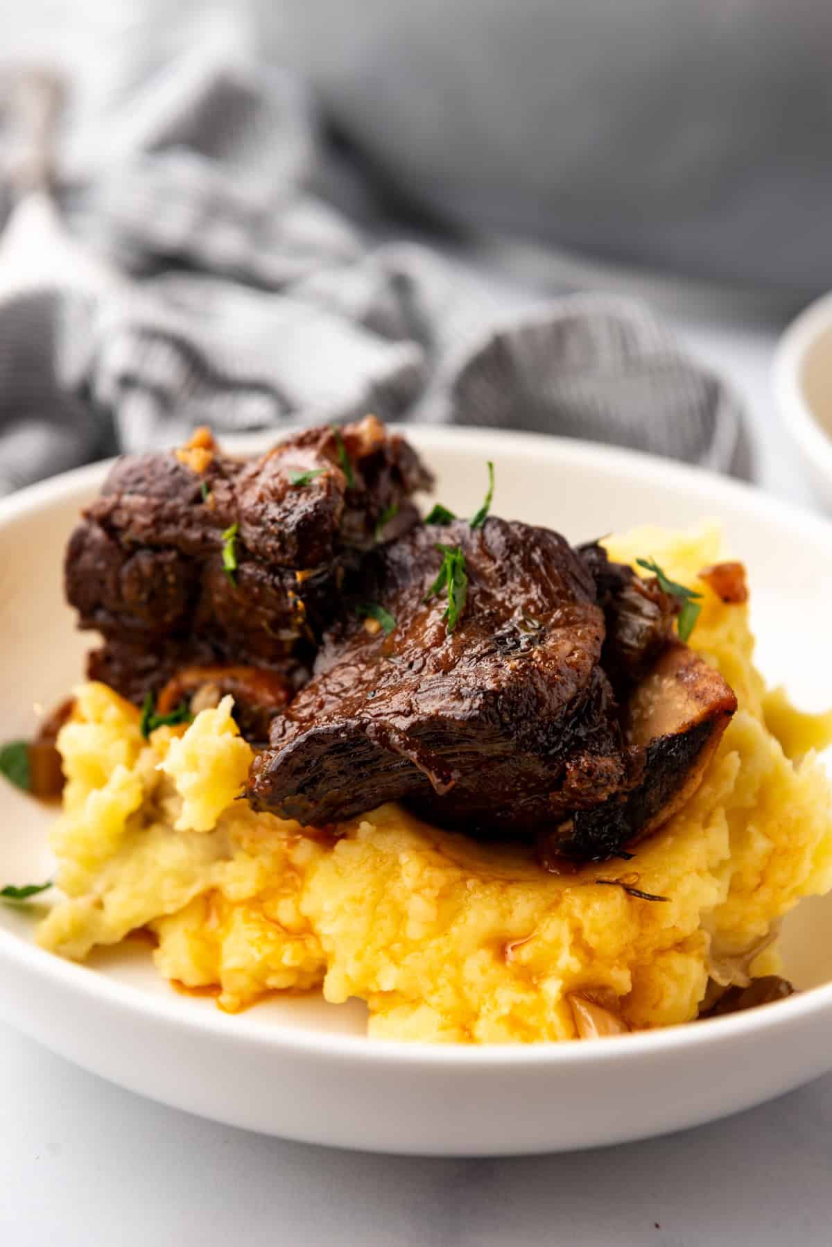 Close up image of a serving of Braised Short Ribs on a bed of mashed potatoes, in a white plate on a white countertop.