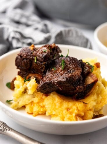 Close up image of a serving of Braised Short Ribs on a bed of mashed potatoes, in a white plate on a white countertop.