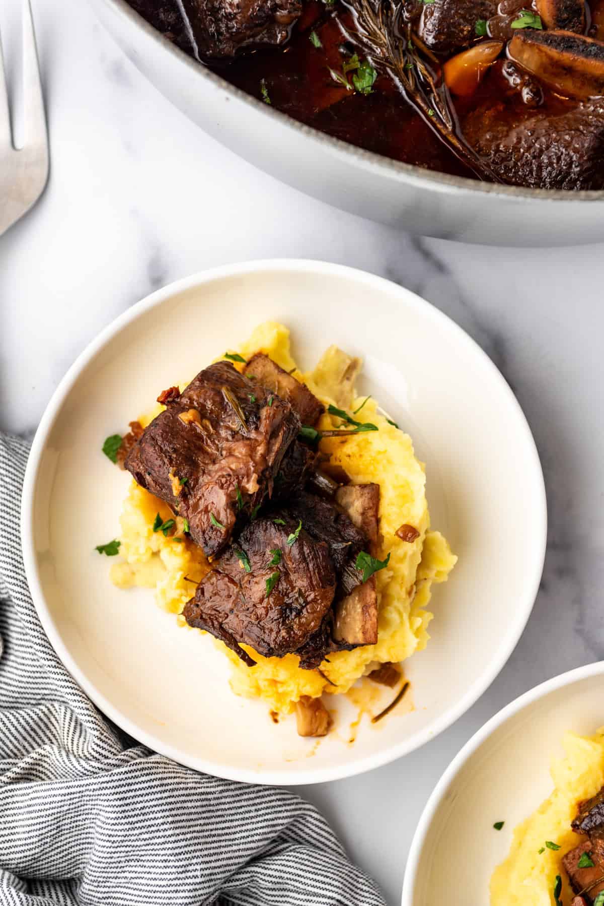 Overhead image of a serving of Braised Short Ribs on a bed of mashed potatoes, in a white plate on a white countertop.