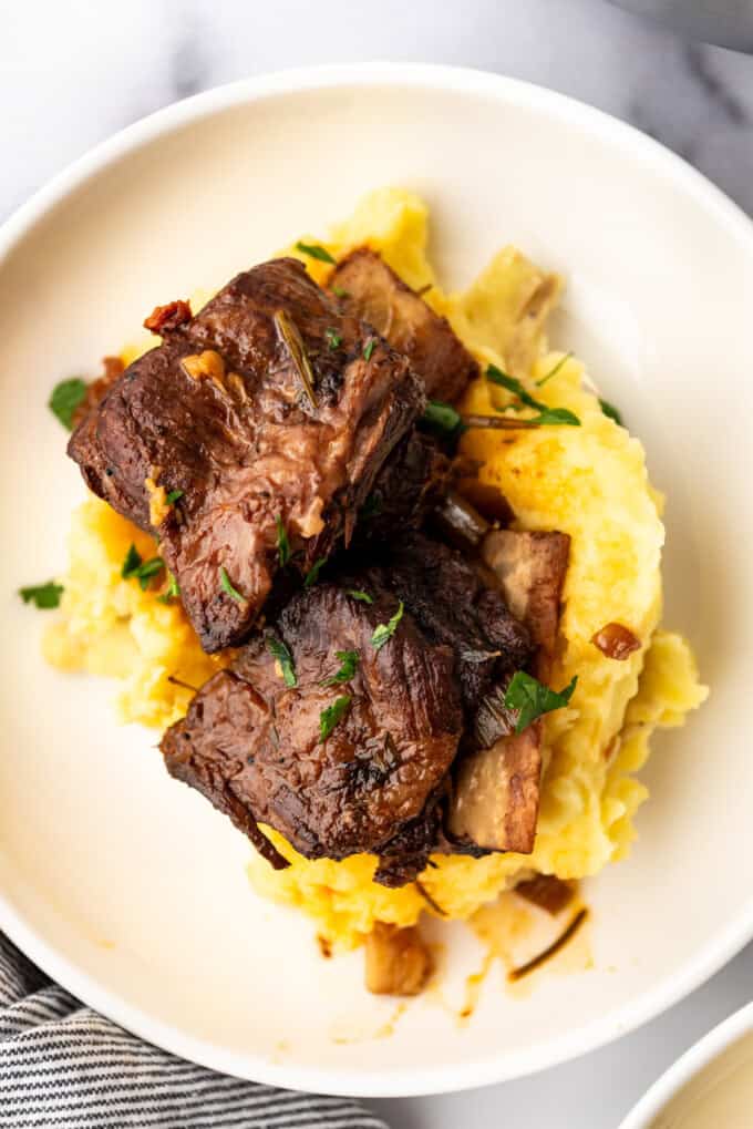 Overhead image of a serving of Braised Short Ribs on a bed of mashed potatoes, in a white plate on a white countertop.