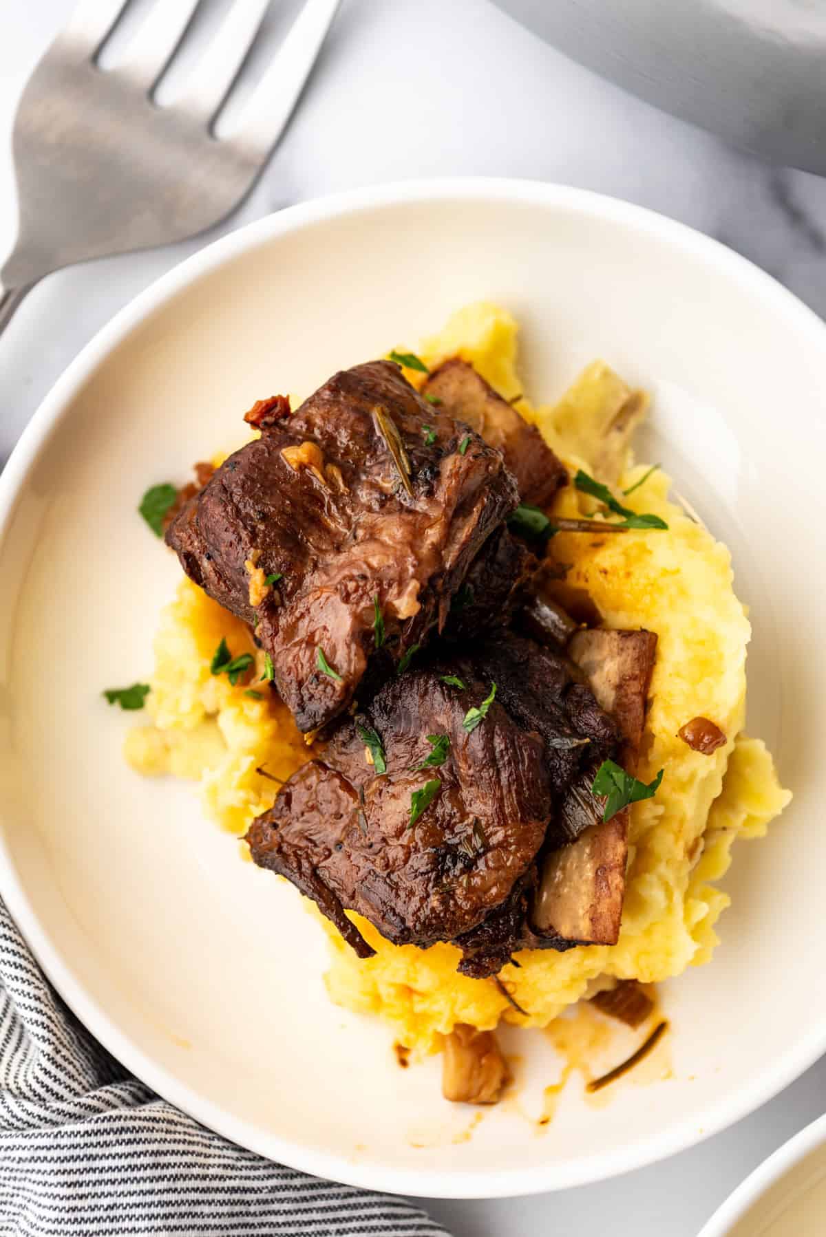 Overhead image of a serving of Braised Short Ribs on a bed of mashed potatoes, in a white plate on a white countertop.