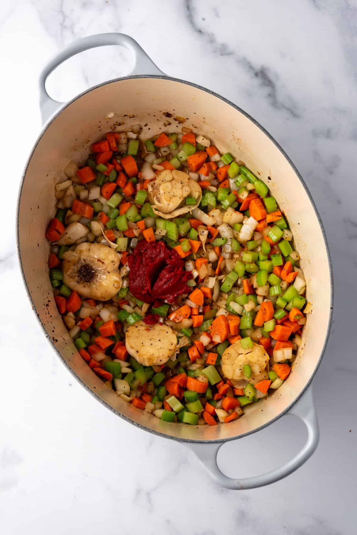 Overhead image of large dutch oven containing garlic, onion, celery, and carrots, with tomato paste on top.