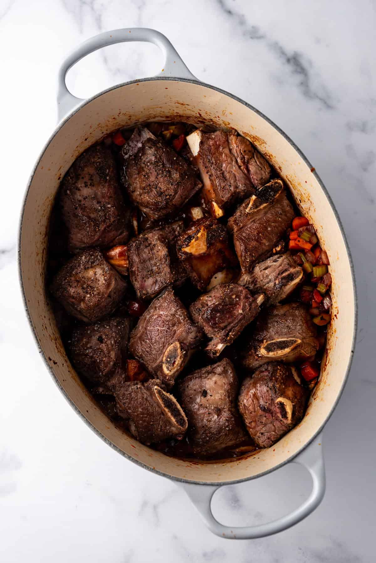 Overhead image of seared short ribs added to large dutch oven with vegetables and sauce.