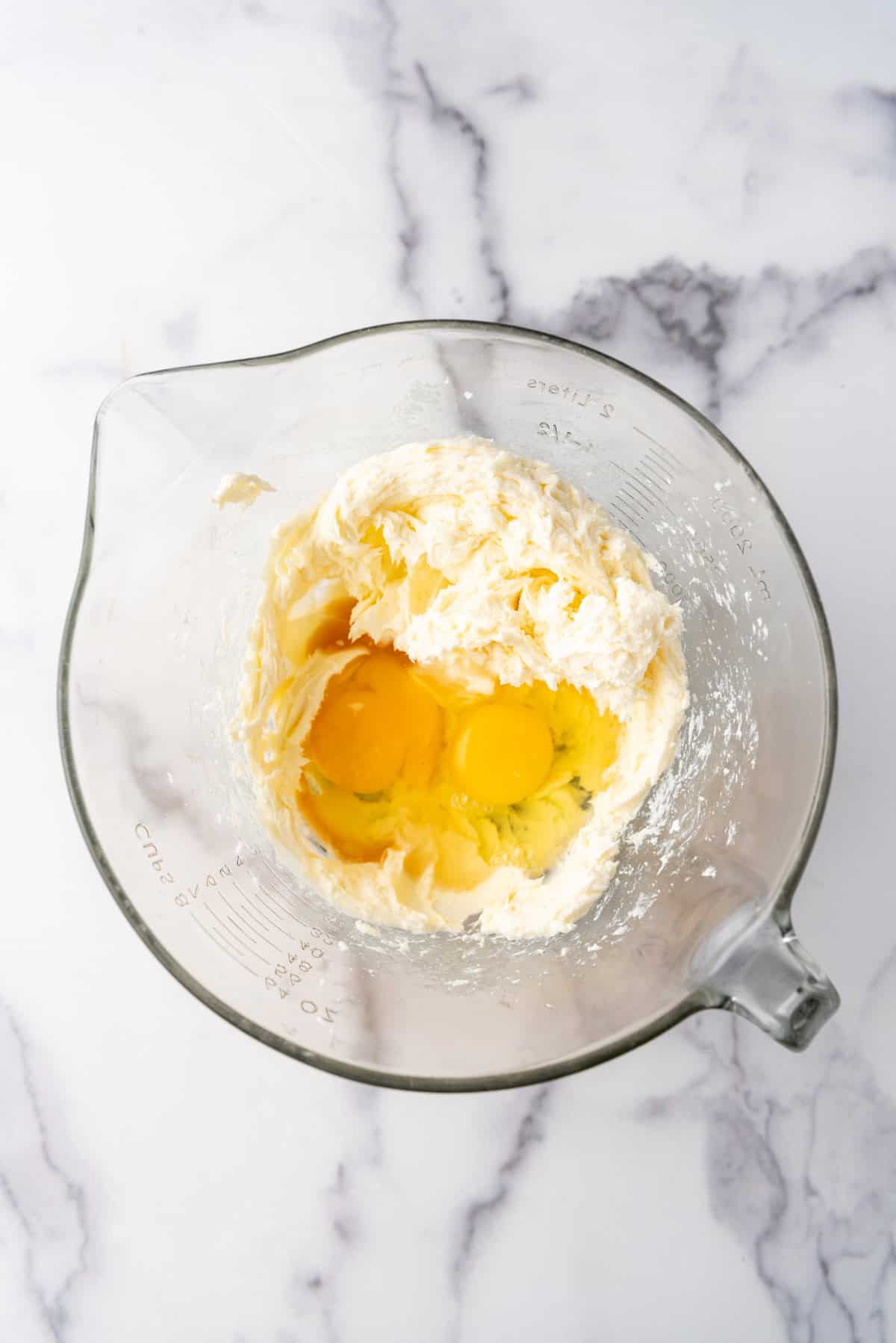 Overhead view of a large glass bowl with eggs and vanilla extract added to a cream cheese mixture.