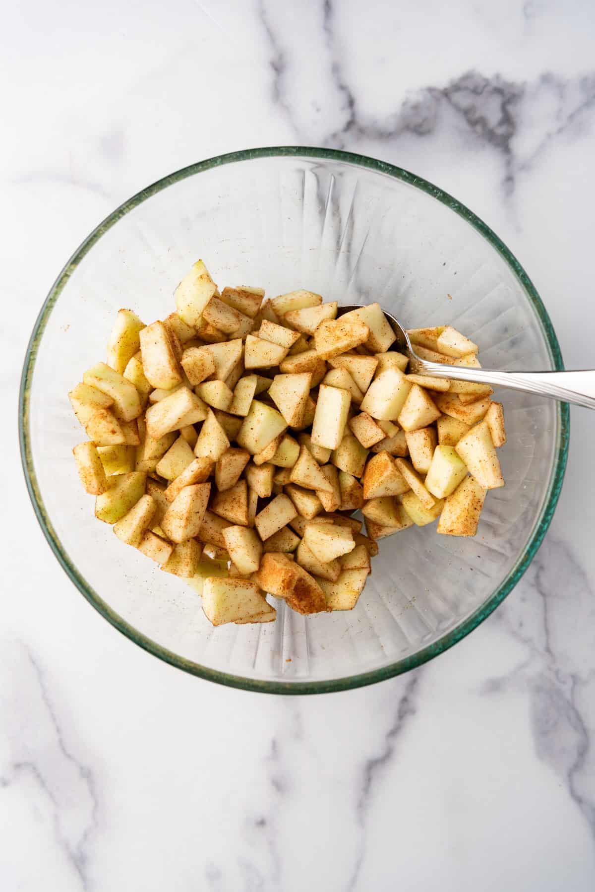 Overhead view of chopped apples, sugar, cinnamon, and nutmeg stirred together in a glass bowl.