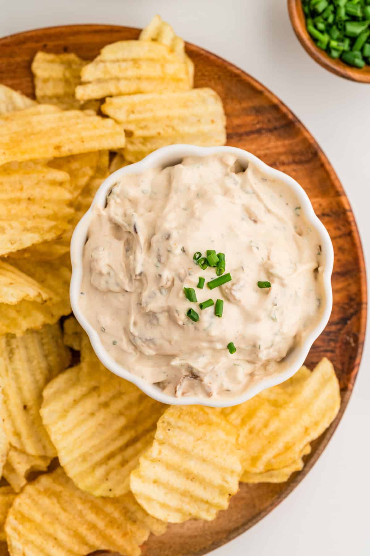 A small bowl of homemade french onion dip on a wooden plate next to potato chips.