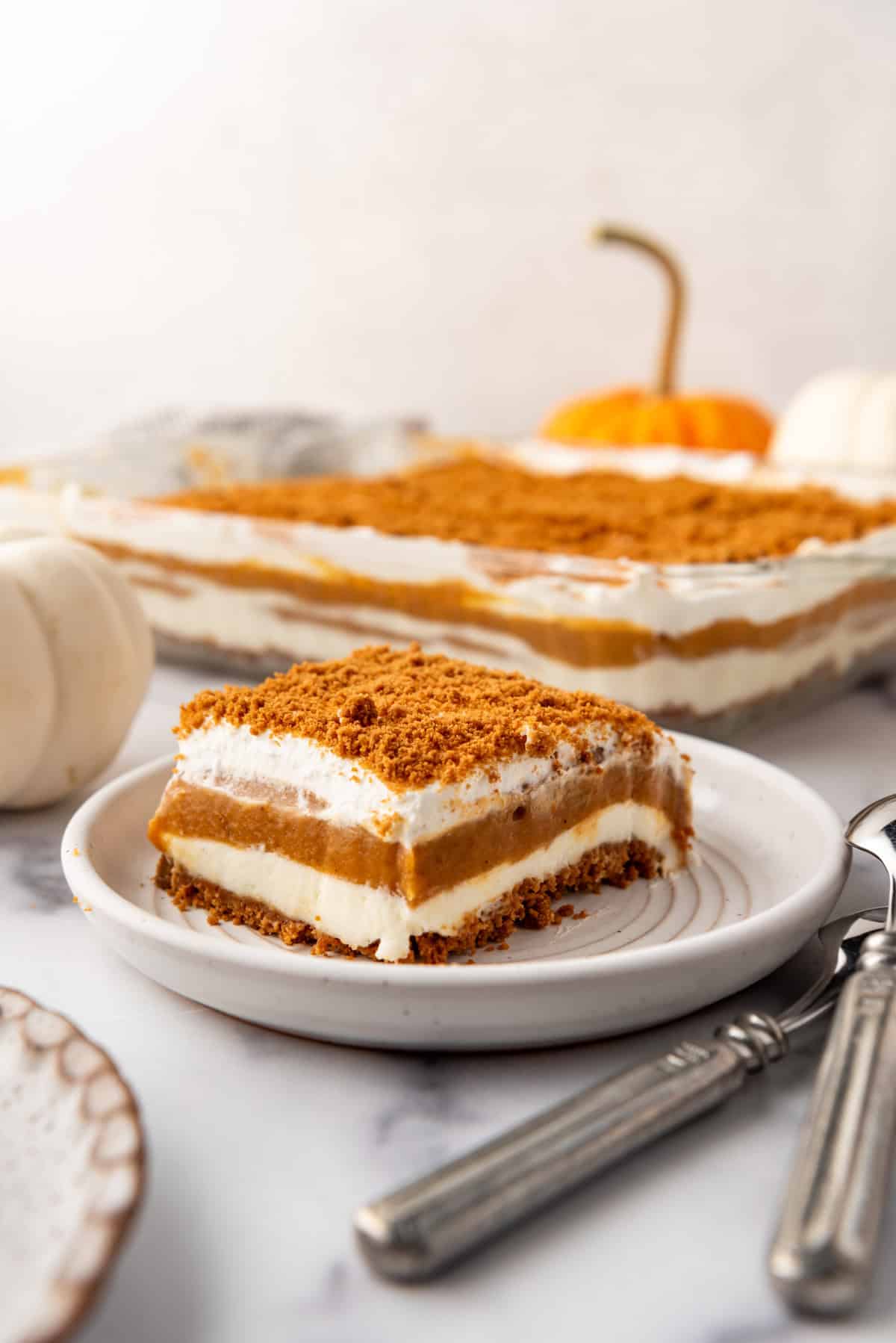 An image of layered no-bake pumpkin lush dessert on a plate next to spoons and a white pumpkin.