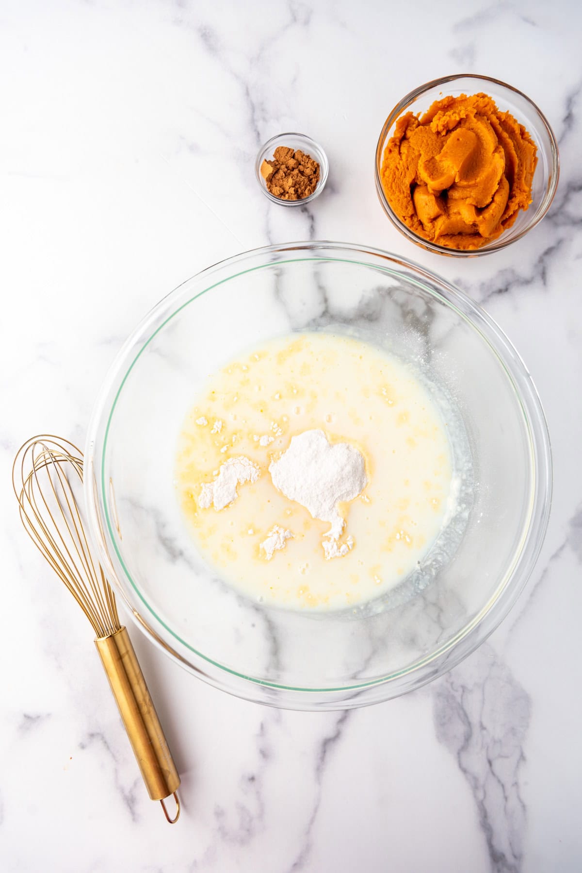 Combining pudding mix and milk in a glass mixing bowl next to a whisk and bowls of pumpkin and spices.