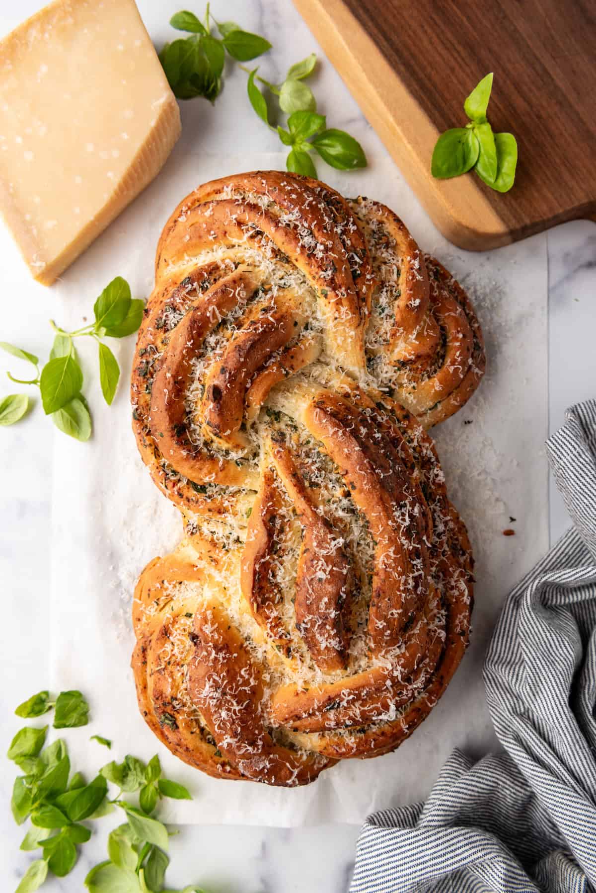 An overhead image of a loaf of swirled garlic herb bread next to fresh basil leaves and a brick of parmesan cheese.