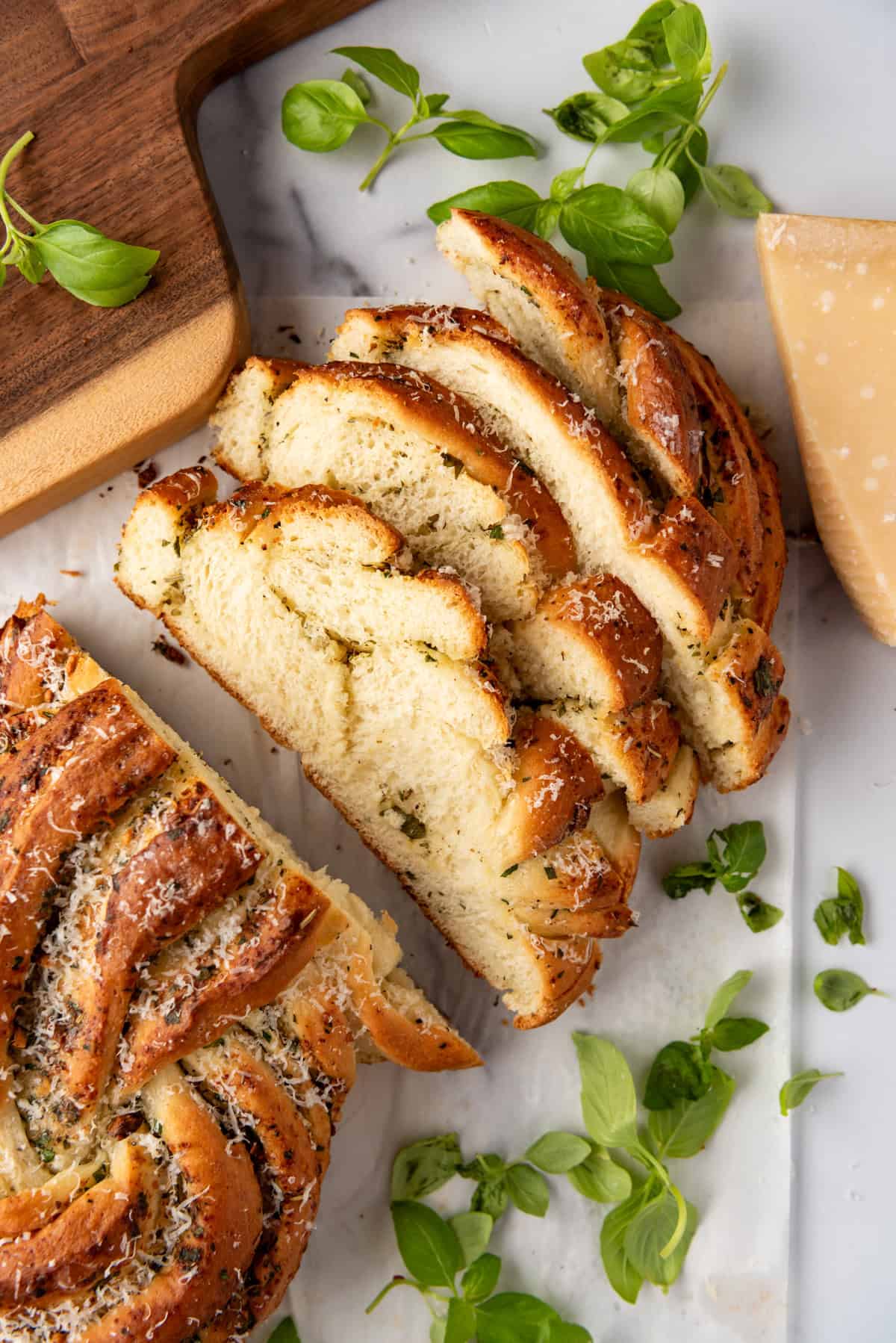 An overhead image of a loaf of sliced bread surrounded by fresh basil leaves, parmesan cheese, and a wooden cutting board.