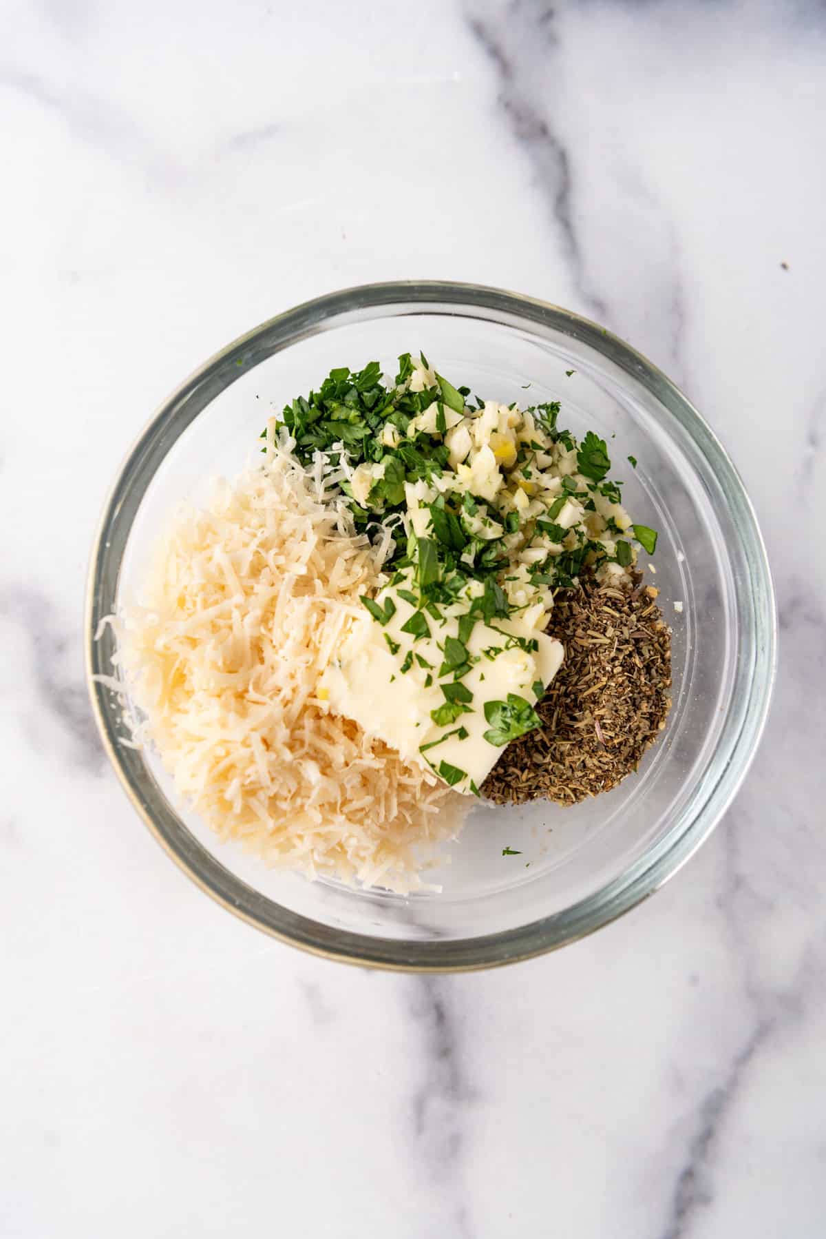 Combining softened butter, parmesan cheese, minced garlic, herbs, and spices in a glass bowl.