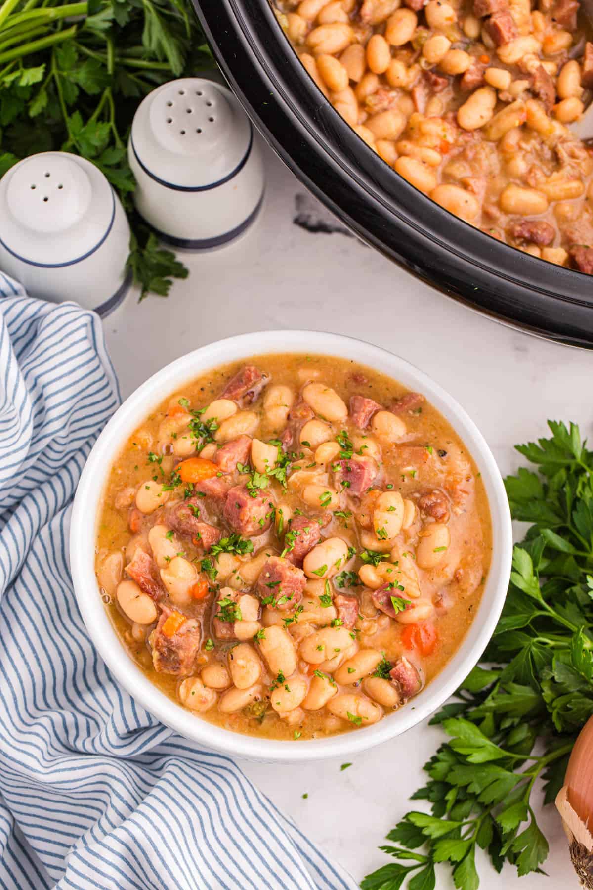 A bowl of crock pot ham and bean soup surrounded by salt and pepper shakers, parsley, and a striped napkin.