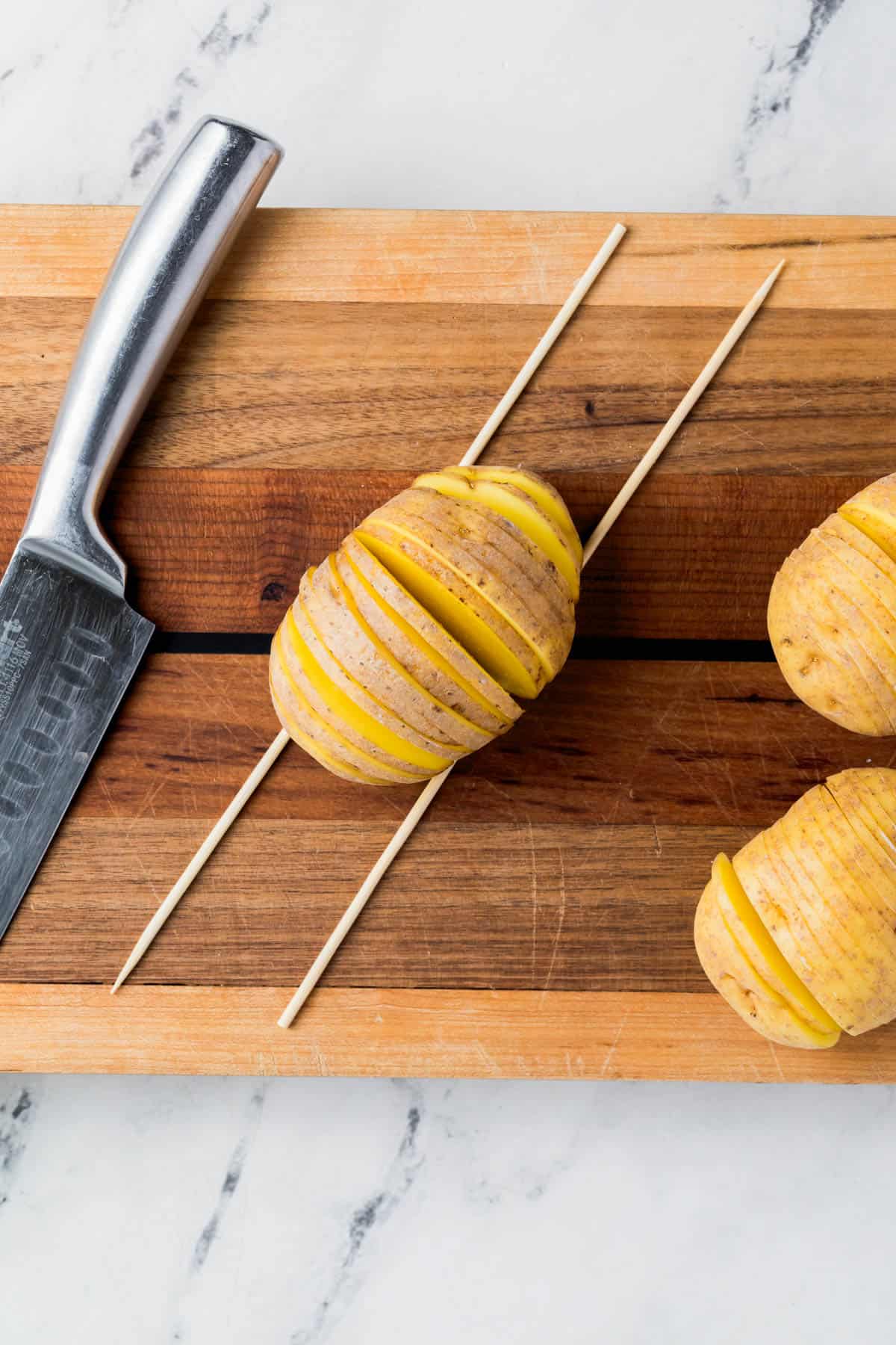 A potato between two chopsticks on a cutting board next to a knife.