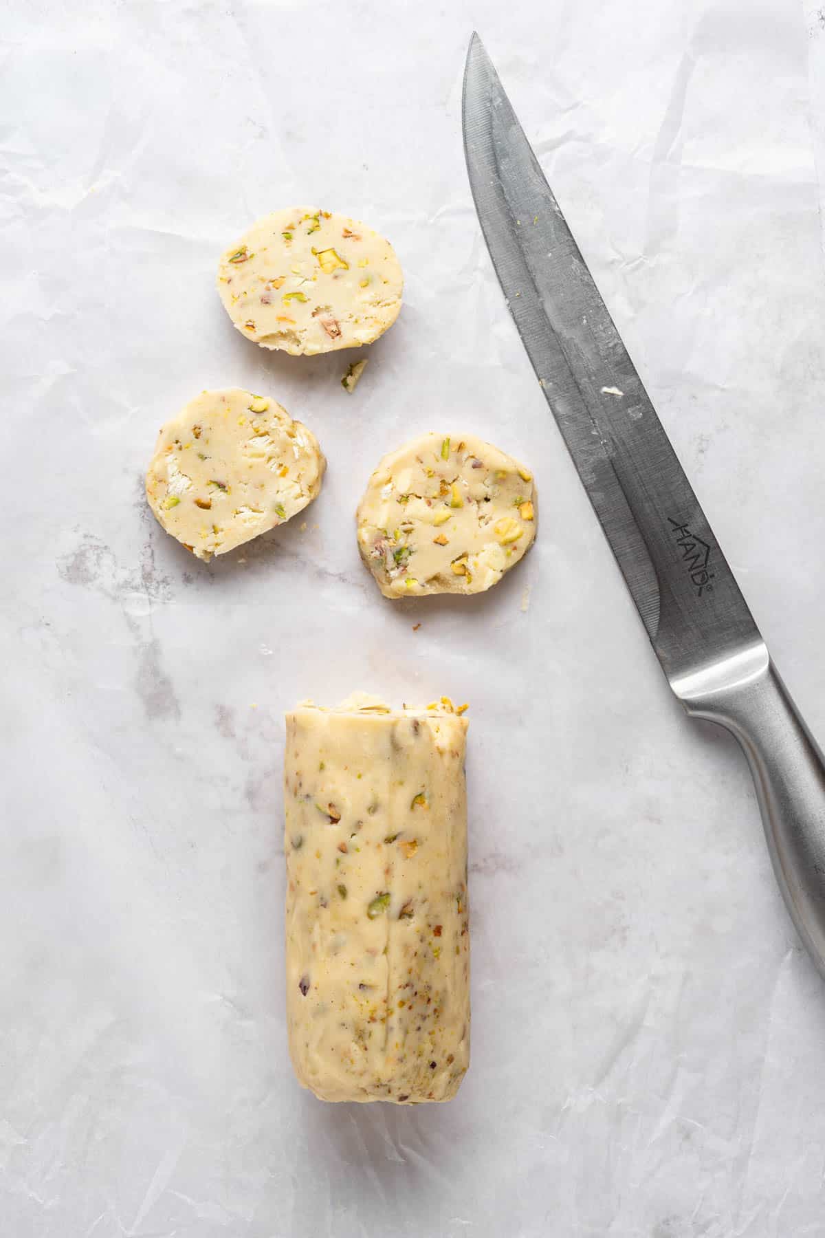 Overhead view of a log of Pistachio Shortbread cookie dough being sliced into individual cookies on a white surface.